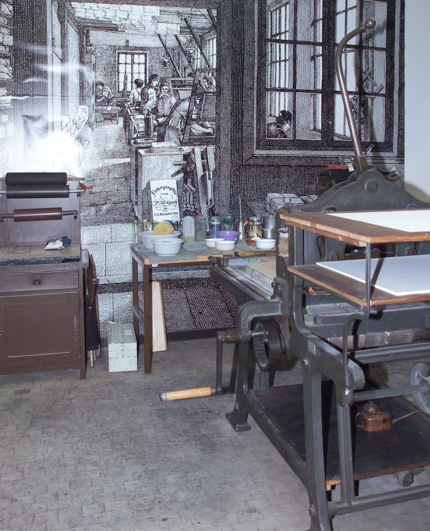 Eine Handhebelpresse steht in einer Druckwerkstatt. Im Hintergrund sieht man weitere Hilfsmittel für den Druck.